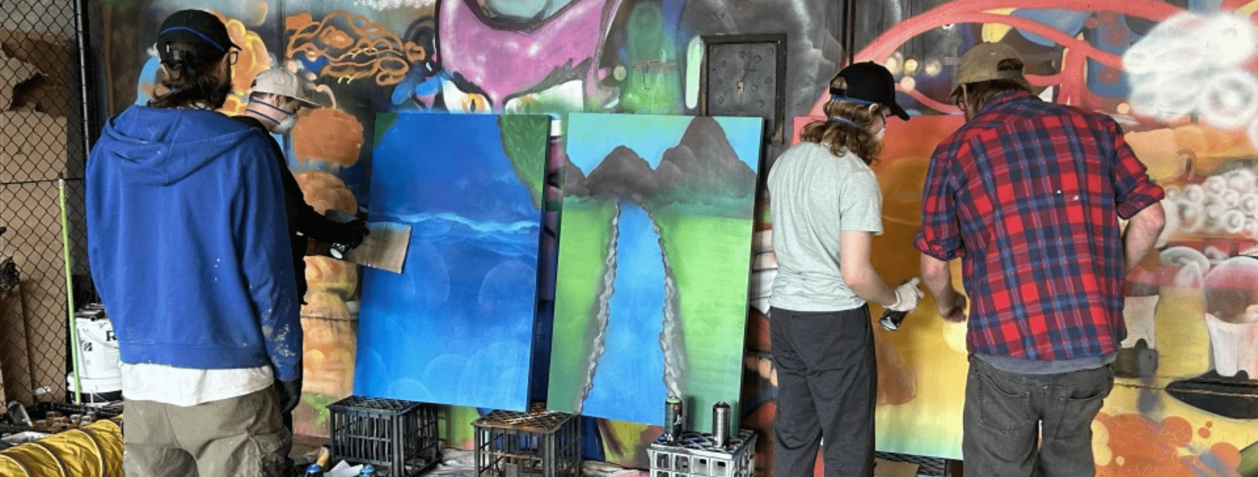 A group of youth residents are guided by a professional graffiti artist on creating artwork. Two young people are spray-painting. Each canvas represents an element: water, air, and earth.
