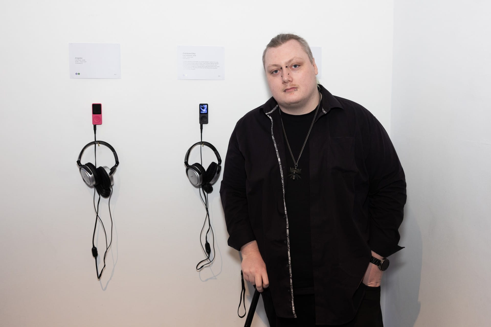 Ciel stands next to a wall at the What Matters Art Expo. Behind him, headphones and MP3 players hang on the wall,  display text reads: Overhead Boy, Ciel.