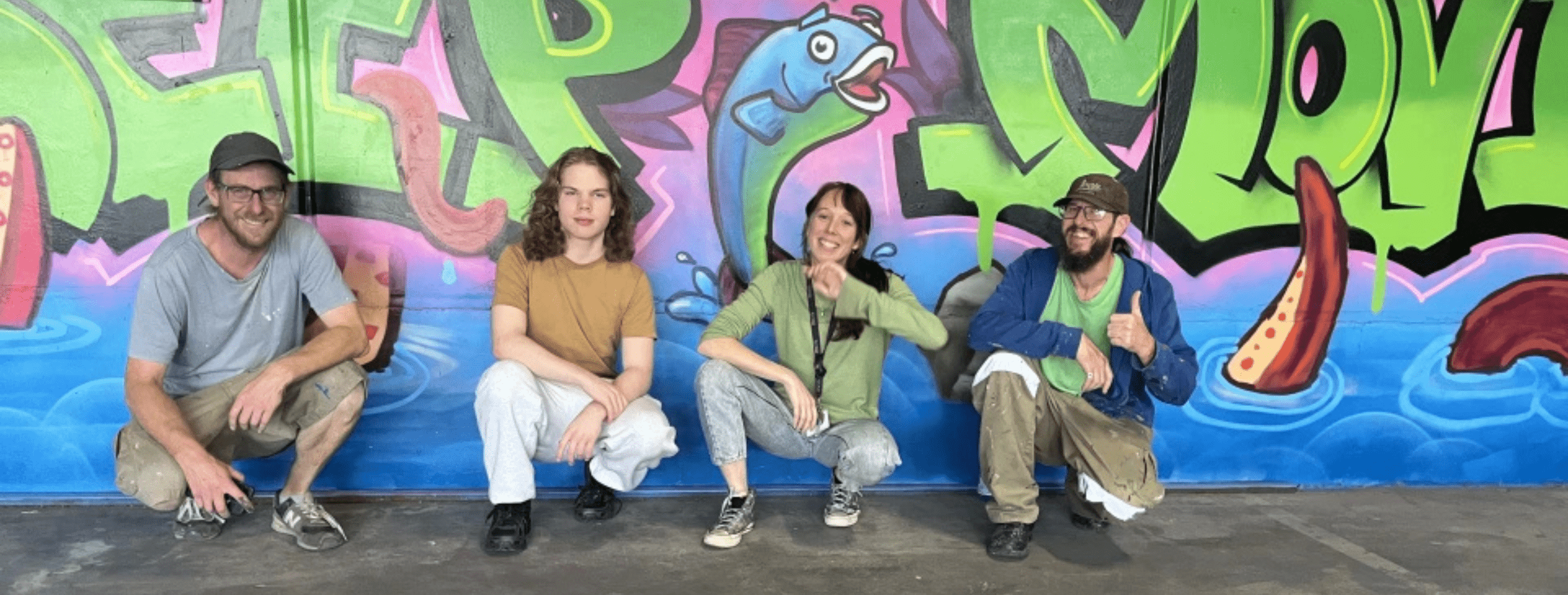 Four people crouch in front of a graffiti mural. The mural reads 'Keep moving'.  