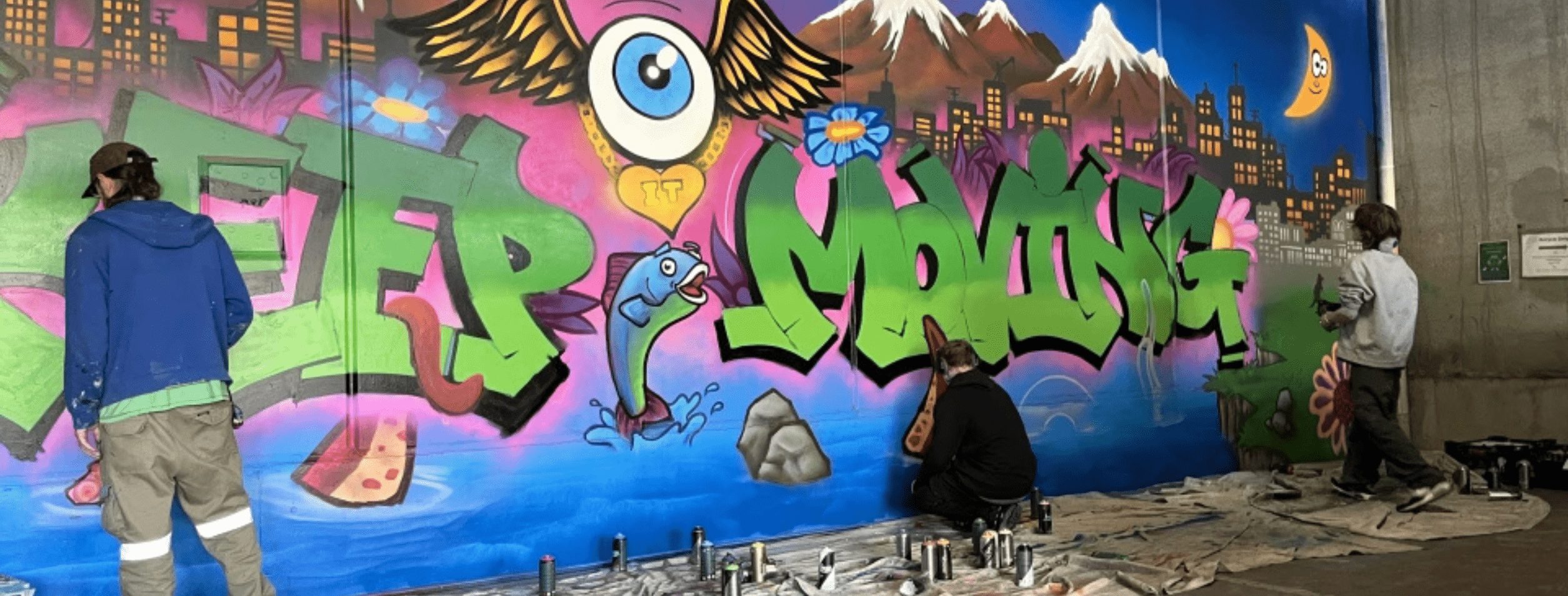 Three youth residents are spray-painting a wall. The graffiti artwork is colourful with a vivid blue, pink and purple gradient. Text reads "Keep it moving."