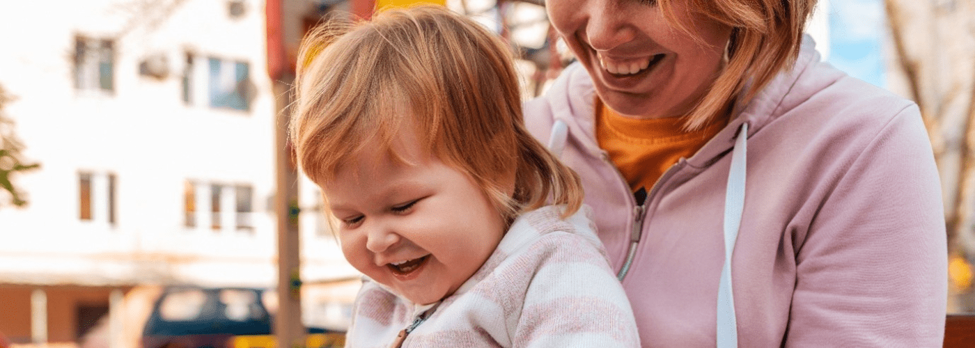A parent holds a child in their lap, the parent is reading a book to them as the child laughs.