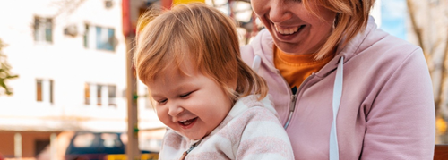A parent holds a child in their lap, the parent is reading a book to them as the child laughs.