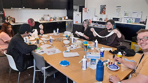 A group of young teenagers sit around a kitchen table eating and participating in arts and crafts activities