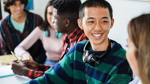 A teenage boy with black hair and ear phones placed around his neck, talks to a female teenager in a classroom setting