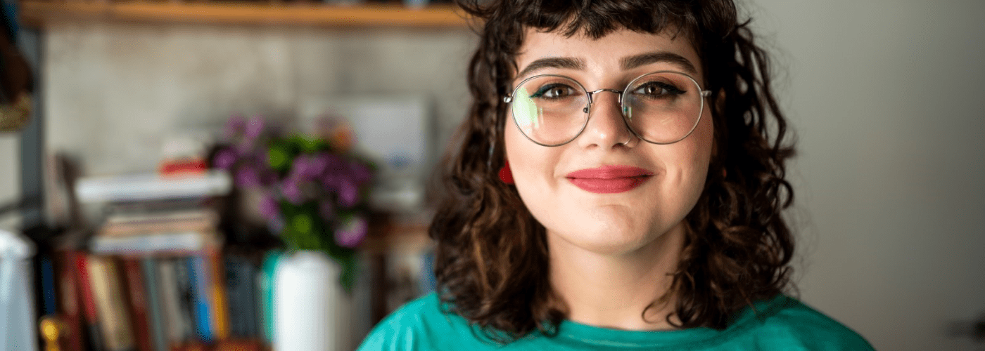 Woman with brown curly hair, large round glasses, red lipstick and a green jumper smiling at the camera. Photo is cropped to her face and shoulders. In the background there is a bookshelf.
