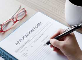 photo of application form on a table with a pair of red eye glasses, a cup of coffee and a hand holding a pen