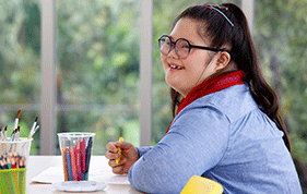 Teenage girl with glasses and black hair sits at the table colouring