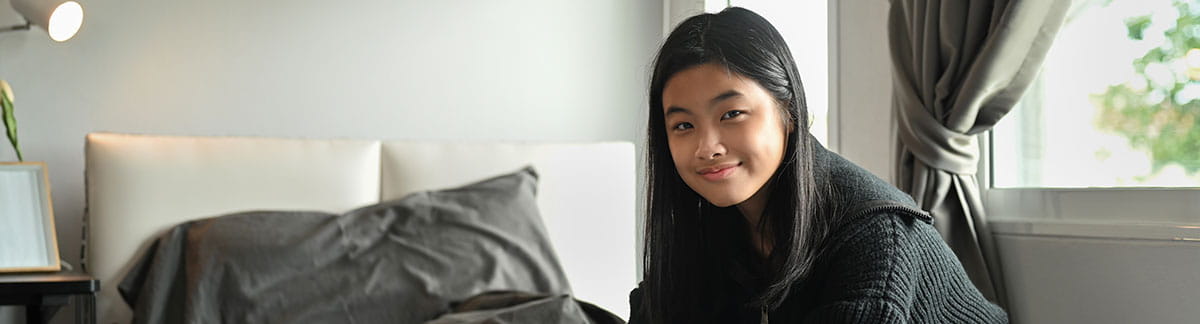 Young female teenager with straight long black hair sitting on her bed