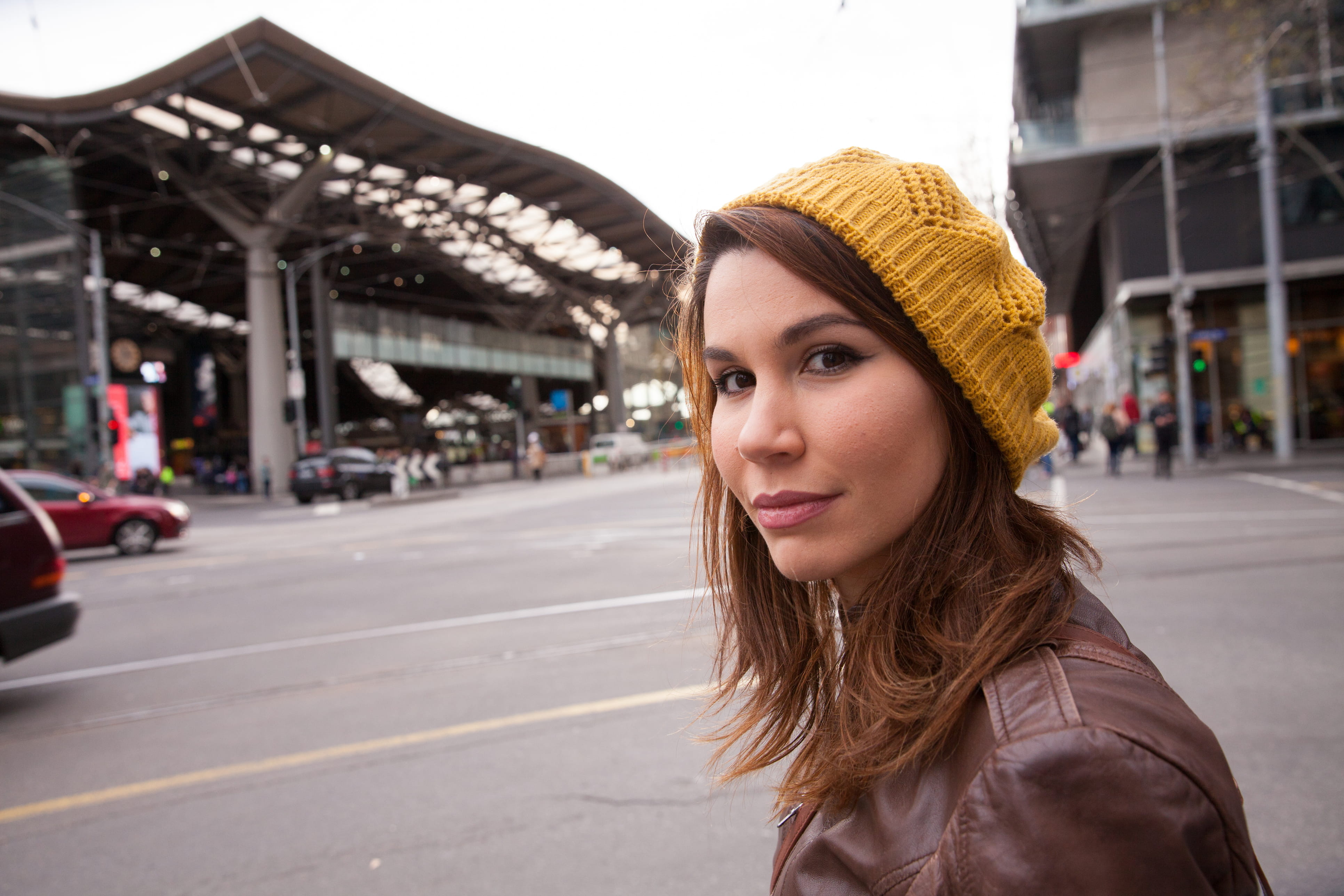 Young woman wearing a yellow knitted beanie and brown leather jacket standing at Spencer Street intersection, with a modern architectural structure and street traffic in the background. She looks confidently at the camera.
