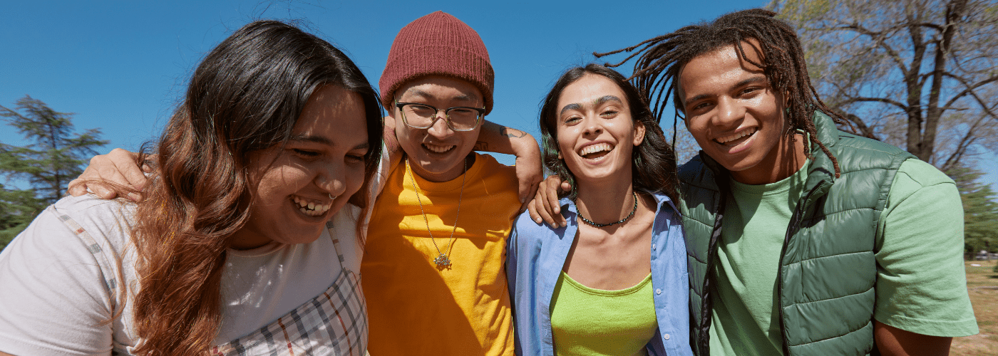 A group of four diverse young adults standing close together outdoors, smiling and laughing under a clear blue sky, showing connection and joy.