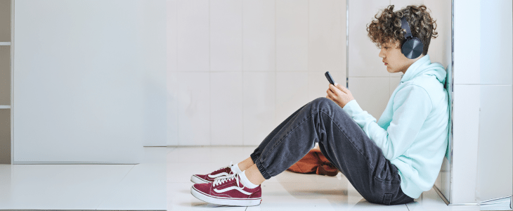 Teenage boy with brown curly hair and big headphones wearing a pale blue hoodie, jeans and red sneakers. Sitting on the floor watching their phone. An orange bag is beside him. 