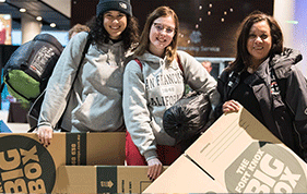 Three females wearing beanies and holding cardboard boxes at Sleep At The G