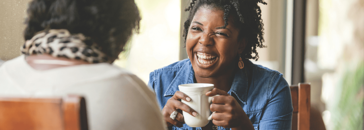 Two women of African descent are sitting and talking, one is wearing a blue shirt and holding a coffee cup facing the camera, and the other is facing away from the camera and wearing a white shirt and an animal print scarf.