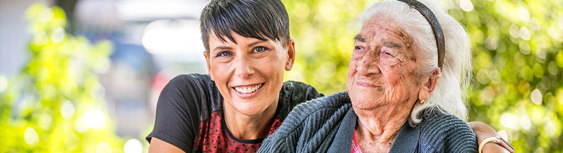 A young female sits and smiles beside an eldely female whyo is also smiling