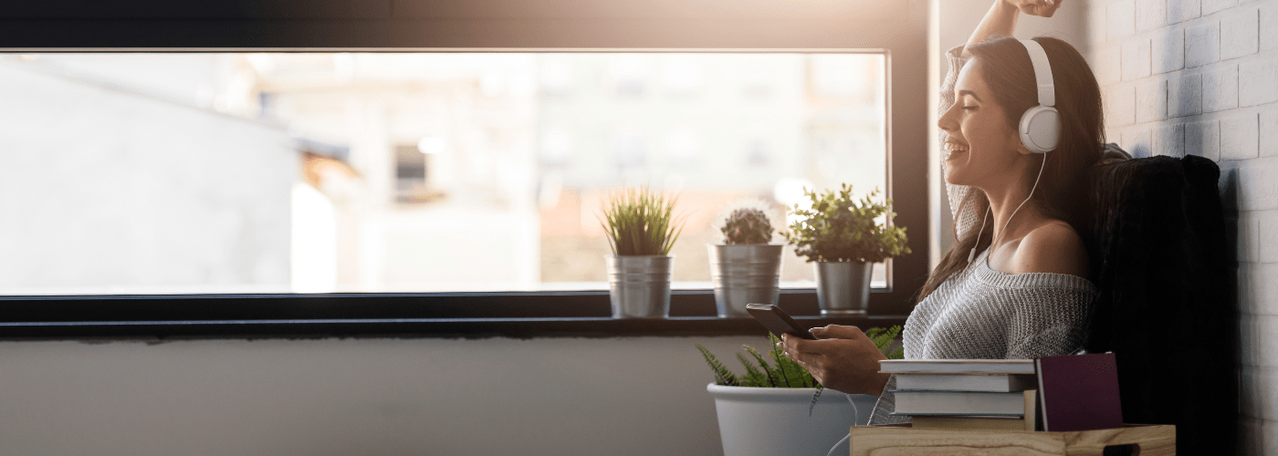 woman sitting at window with headphones on. smiling with one hand up in the air.