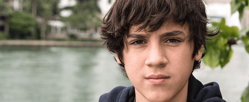 Young teenage boy with brown hair standing on front of a river looking at the camera up close