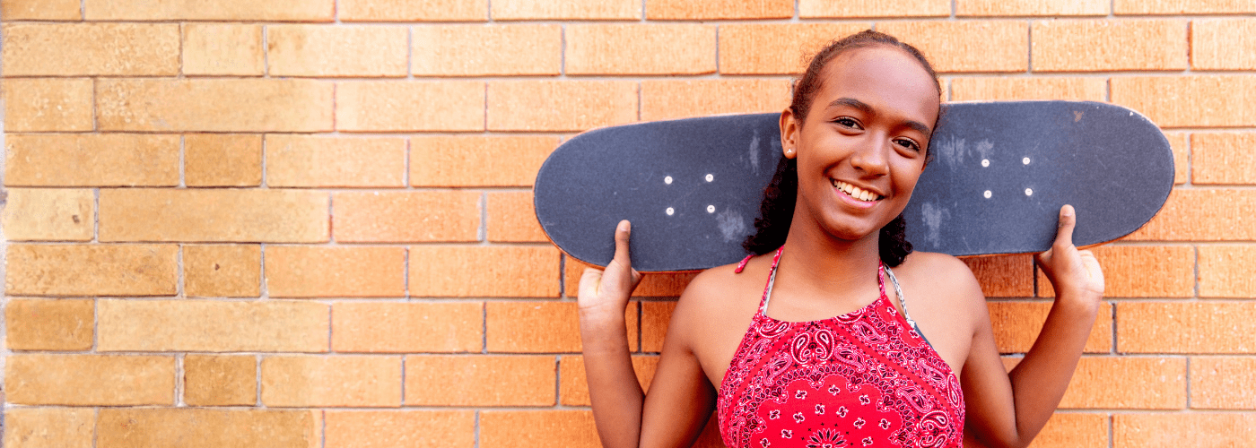 Young Aboriginal teenage girl standing in front of a brick wall holding a skateboard behind her head, smiling