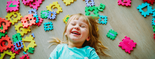 Birdseye photo of a blonde toddler girl laying on the floor with square foam pieces scattered around her - each with letter or numbers cut out from them