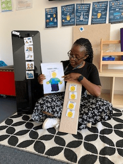  Educator sitting on a patterned rug in a classroom, holding a visual aid that says “Today I feel…” with a picture of a child and emotion symbols. She is also holding a board with emoji-style faces representing different feelings, supporting emotional learning for young children.
