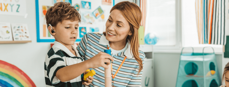 photo of young boy wearing a hearing aid building blocks along side his mother or teacher