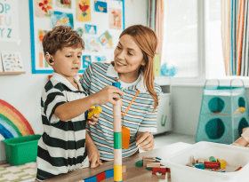 photo of young boy wearing a hearing aid building blocks along side his mother or teacher