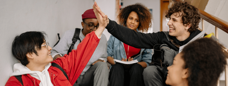 5 teenage students sitting on stairs in a building. 2 of them are reaching across to each other in a high-5.
