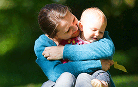 A Mum wearing a blue jumper throws both arms around a baby