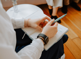 photo focusing on hands resting on a notebook holding a pen. person is sitting opposite a couch where you can see the feet of 2 people sitting