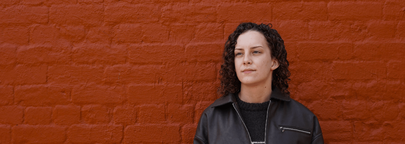 A women wearing a black leather jacket stands against a red brick wall