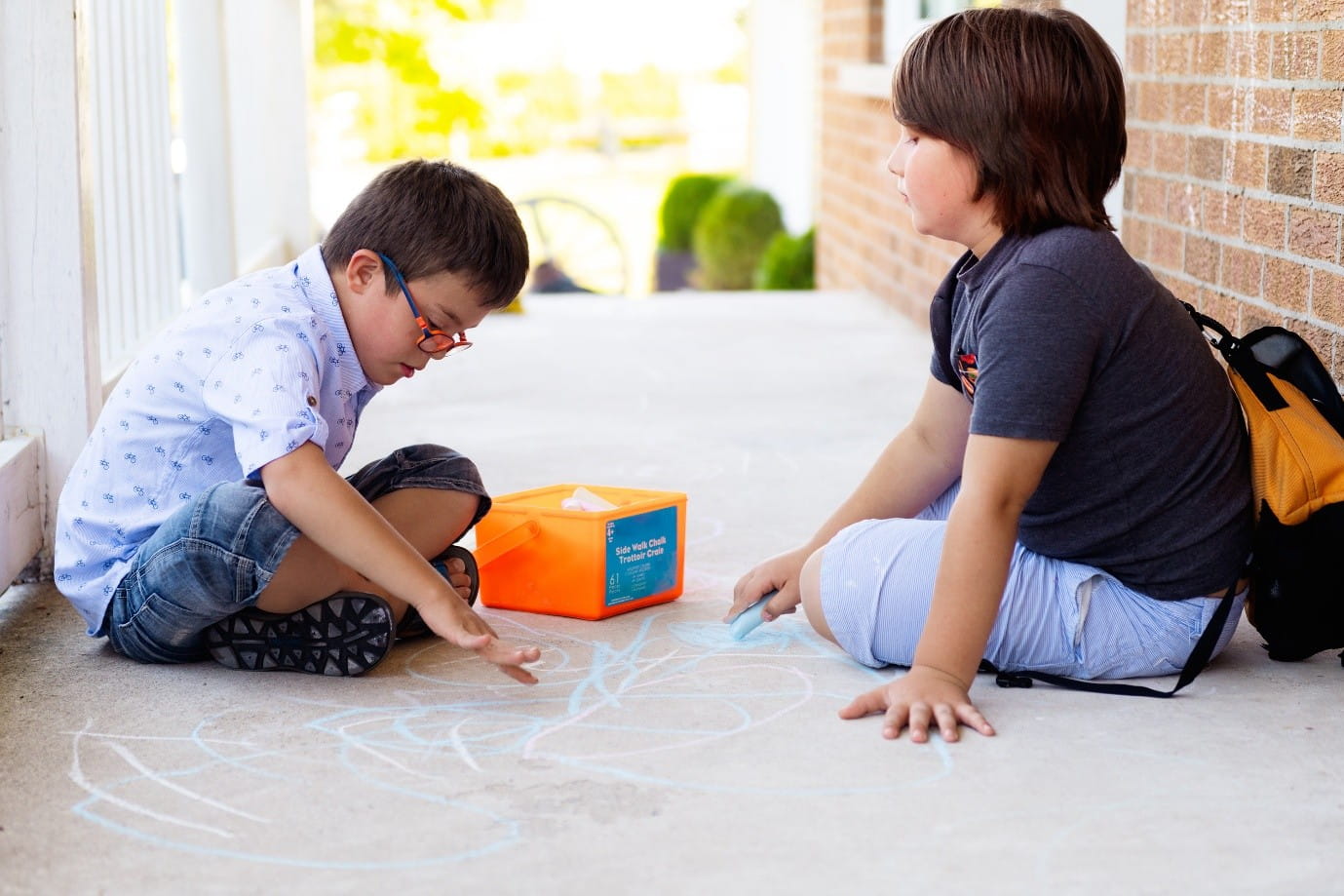 Two young children play together outside, one holds a piece of blue chalk, drawing on the concrete.
